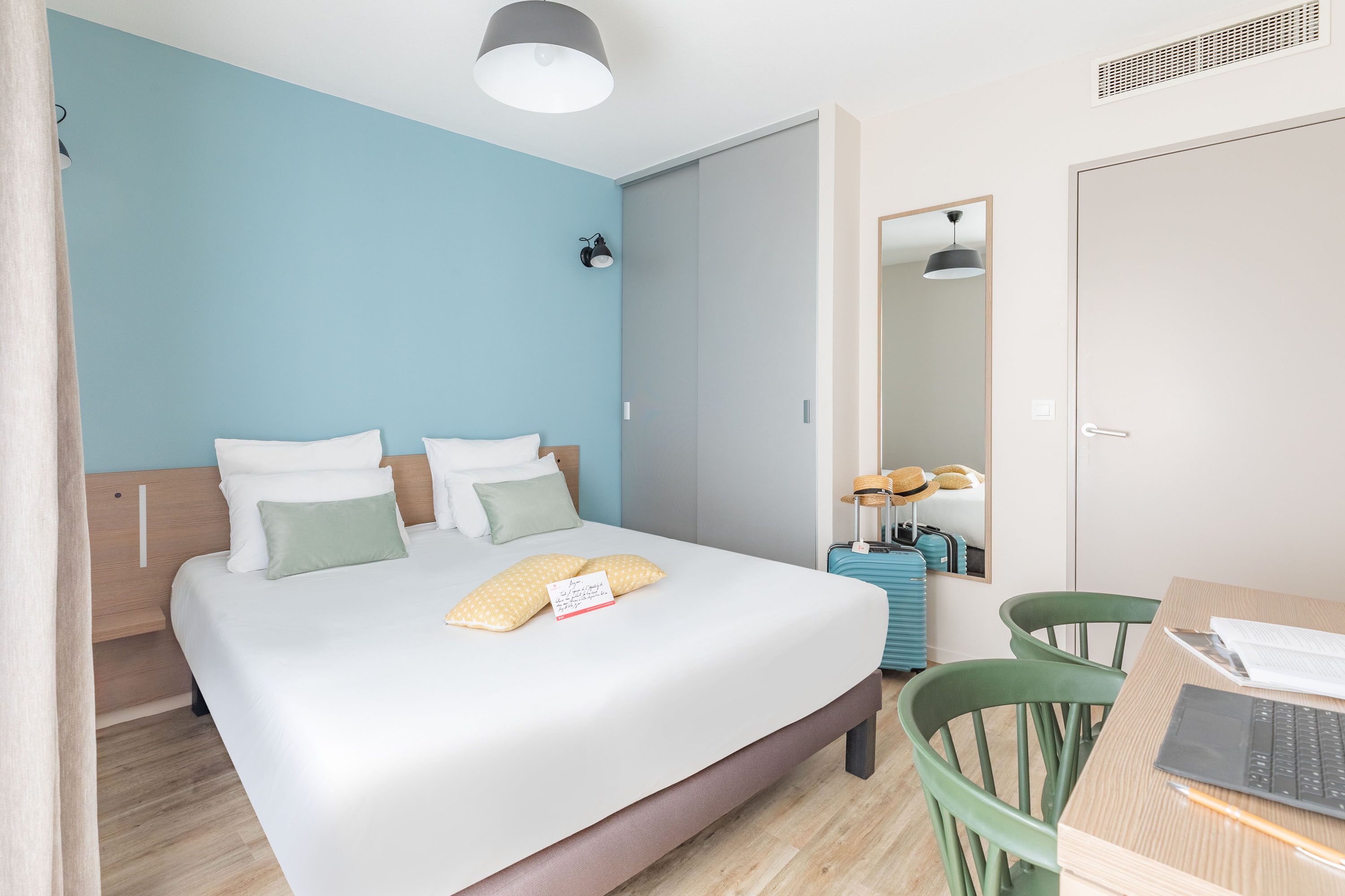 A modern hotel room with a large bed, a desk with a laptop, a suitcase, and a mirror.