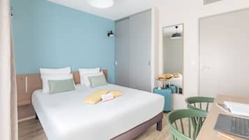 Ein modernes Hotelzimmer mit einem großen Bett, einem Schreibtisch mit Laptop, einem Koffer und einem Spiegel.