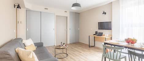 Un salon moderne avec un canapé gris, une petite table ronde, un téléviseur fixé au mur et un coin repas comprenant une table et des chaises en bois.