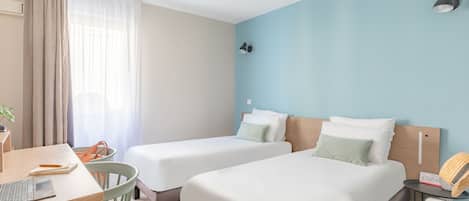 A hotel room with two single beds, a desk with a laptop, a chair, a suitcase, and a lamp.