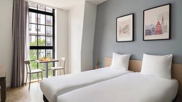 Habitación de hotel con dos camas, una mesa de comedor con sillas, un ventanal con cortinas y dos cuadros enmarcados en la pared.