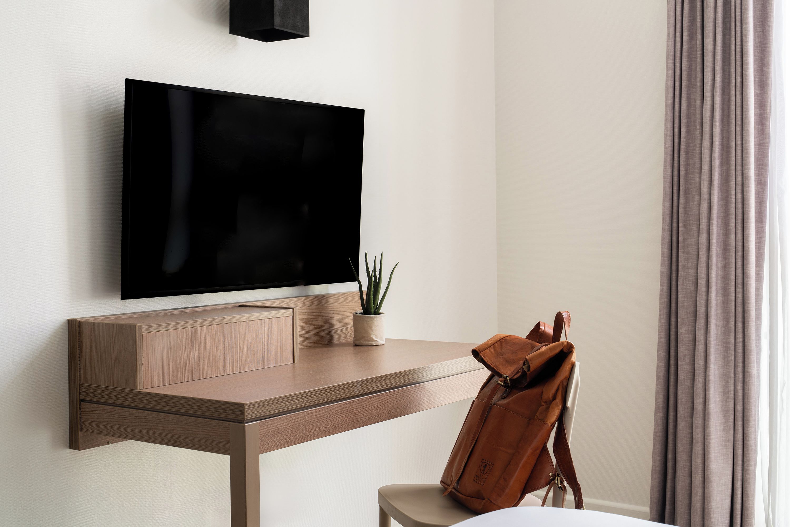 Una habitación moderna con una televisión de pantalla plana en la pared, un escritorio de madera con una planta y un bolso de cuero en una silla.