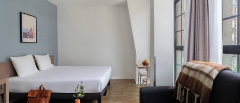 Quarto de hotel moderno com cama, sofá, pequena mesa e janela com cortinas.