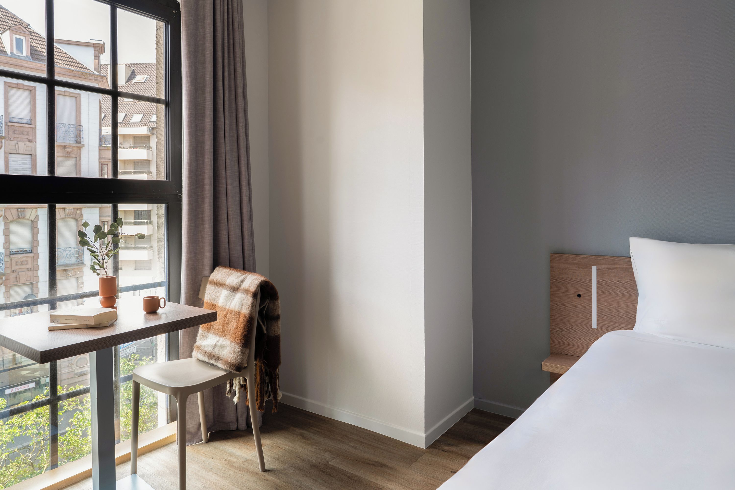 Habitación de hotel con cama, escritorio con silla, ventana con cortinas y vista a edificios.