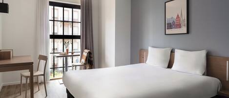 Une chambre d’hôtel avec un lit, un bureau, une chaise, une fenêtre avec des rideaux et un tableau encadré au mur.