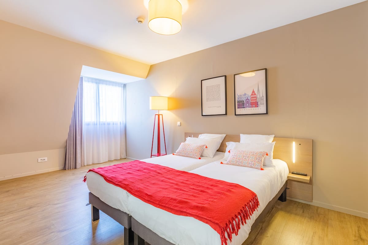 Une chambre d’hôtel moderne avec un grand lit, deux tables de chevet, une lampe sur pied et une fenêtre avec des rideaux.