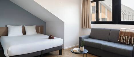 A modern bedroom with a bed, a sofa, a small table, and a window with curtains.