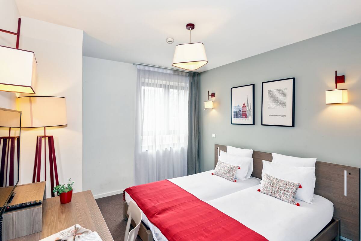 Une chambre d’hôtel avec un lit, un bureau avec un ordinateur, une chaise, une plante rouge et une fenêtre avec des rideaux.