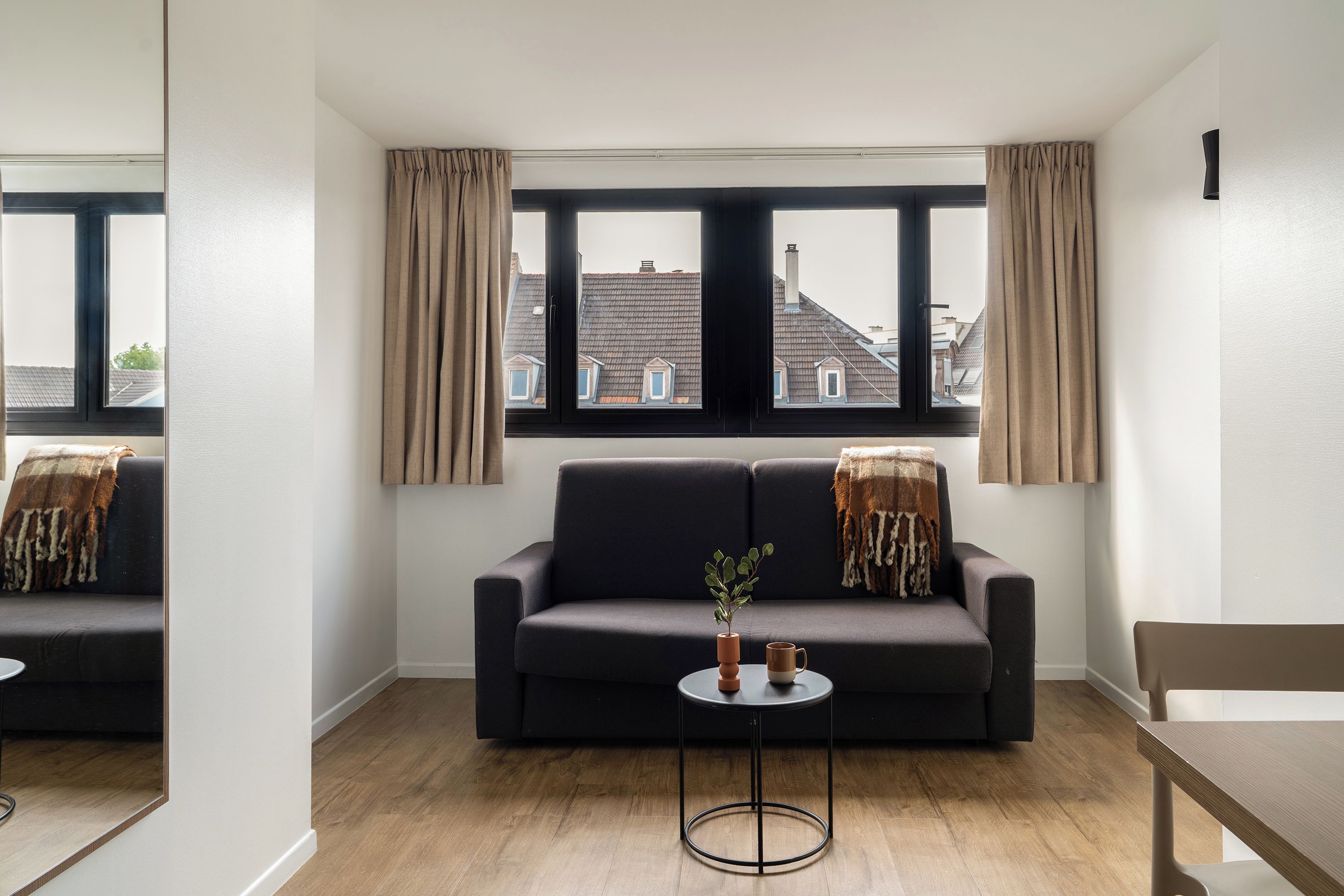 A modern living room with a dark sofa, a small round table with a plant and two cups, and a view of neighboring buildings through large windows with curtains.