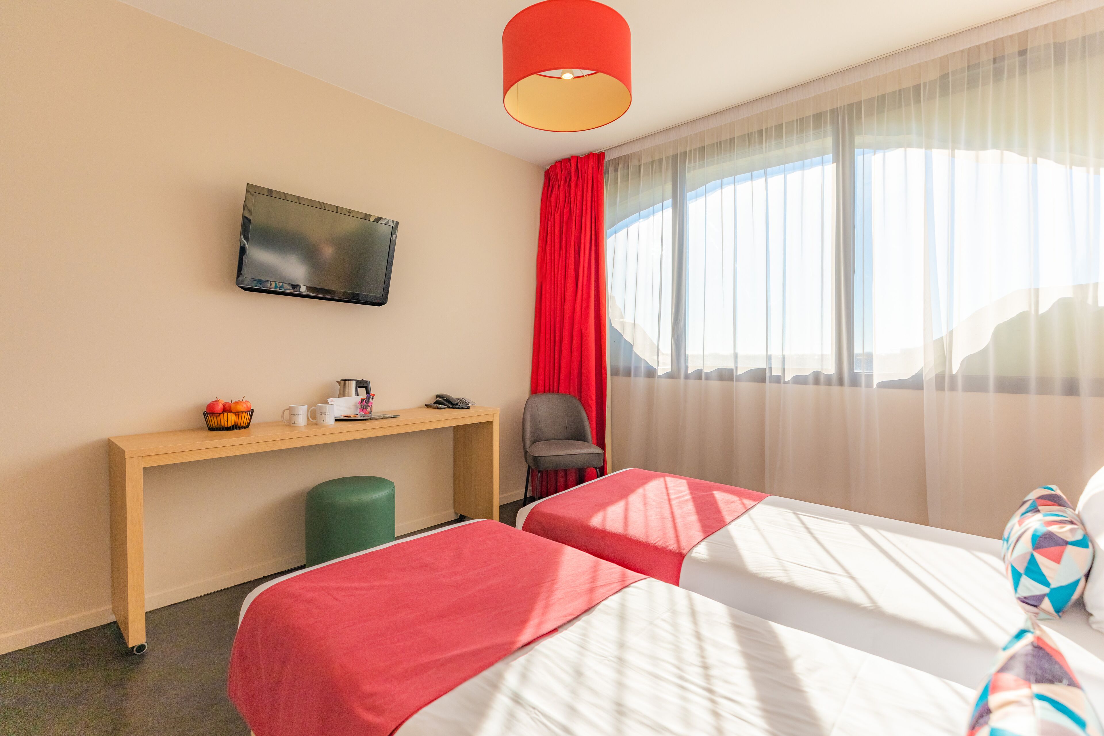Habitación de hotel con dos camas, un escritorio con televisor de pantalla plana, una silla y una ventana con cortinas traslúcidas.