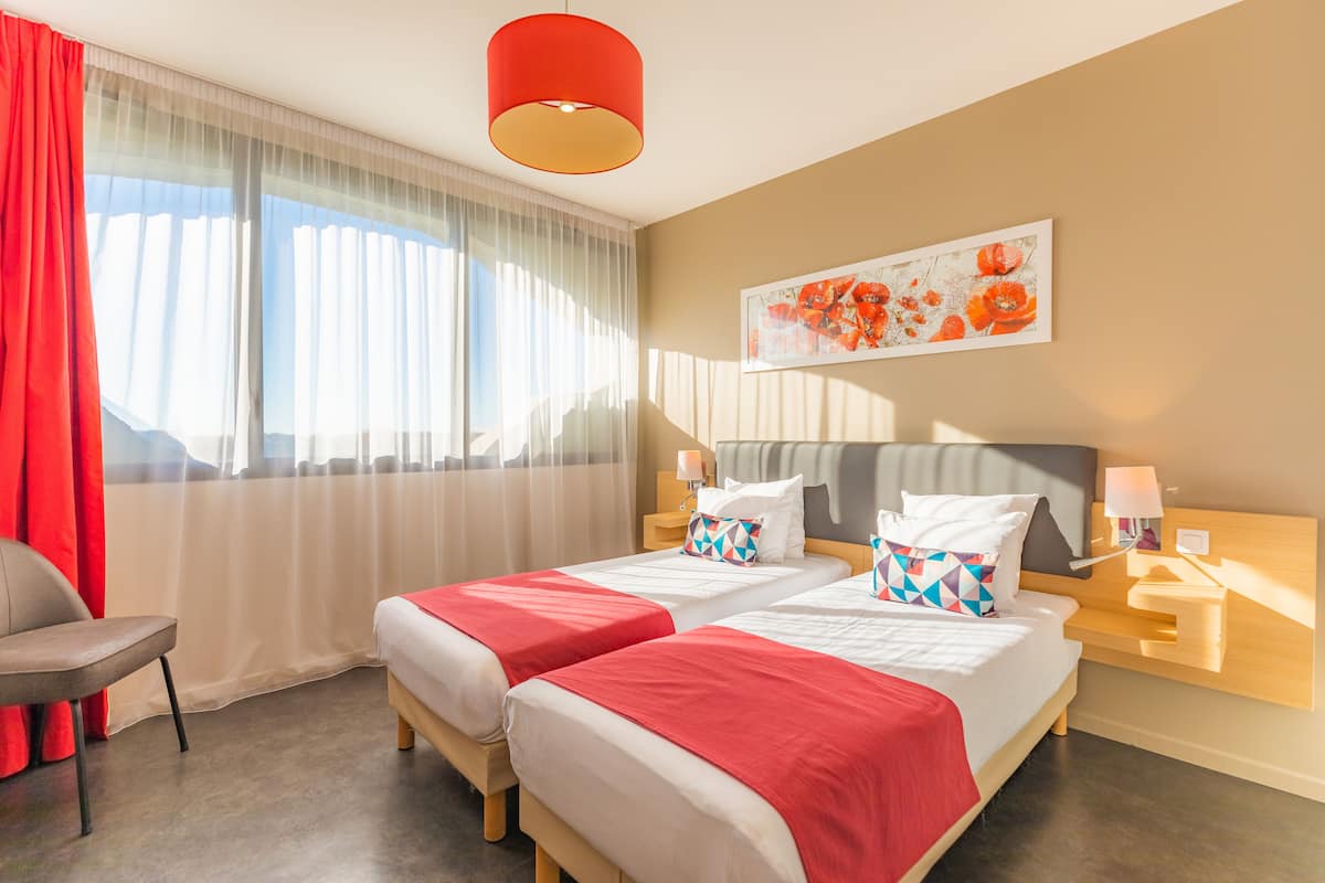 Habitación de hotel con dos camas, una silla roja, un ventanal con cortinas translúcidas, un cuadro en la pared y una lámpara colgante roja.