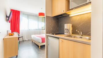 Une chambre d’hôtel compacte avec une kitchenette, un lit agrémenté de rideaux rouges, un petit bureau et une fenêtre avec des rideaux légers.