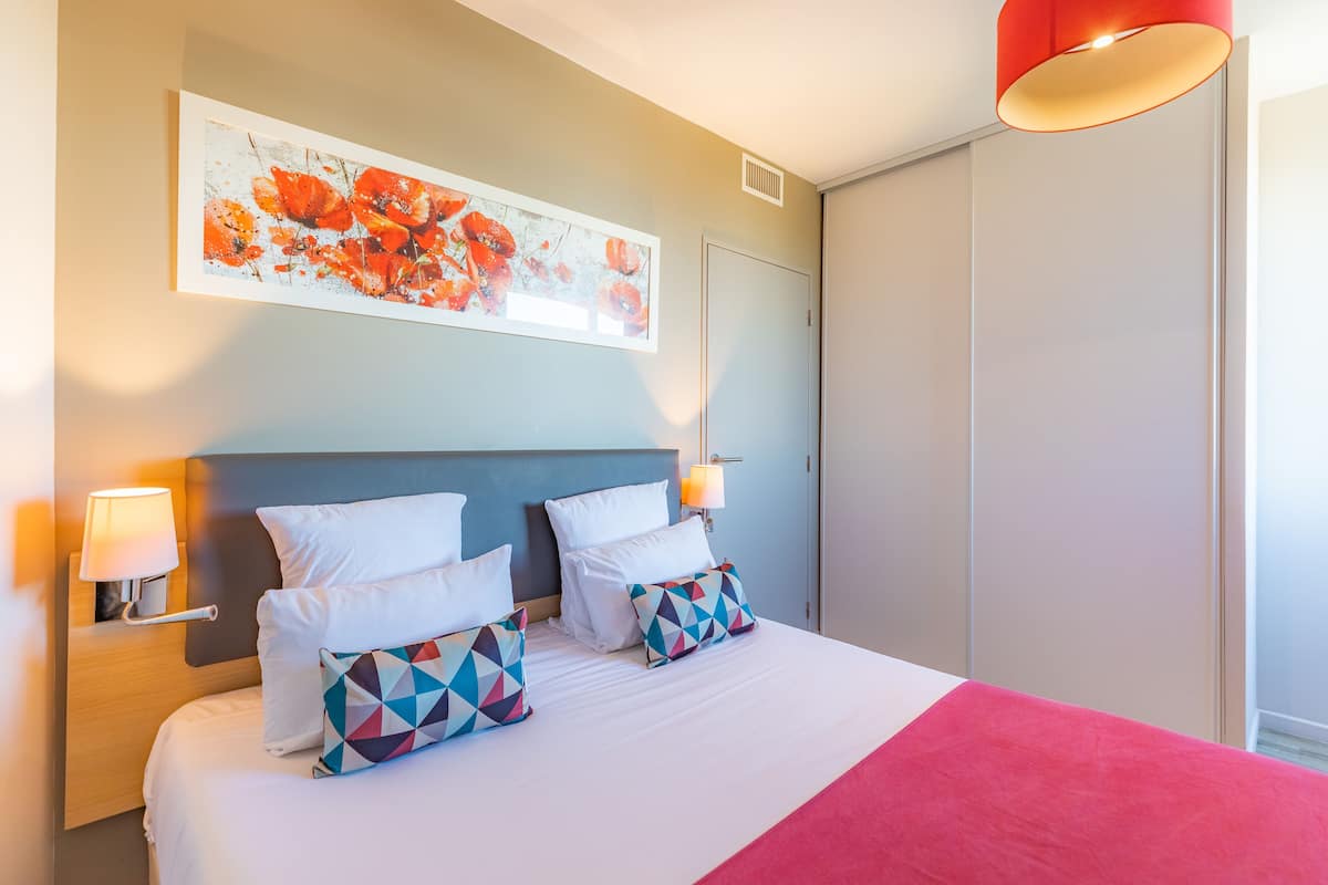 Una cama bien hecha con sábanas blancas y almohadas geométricas de colores, un cabecero azul y un detalle rojo en la colcha.