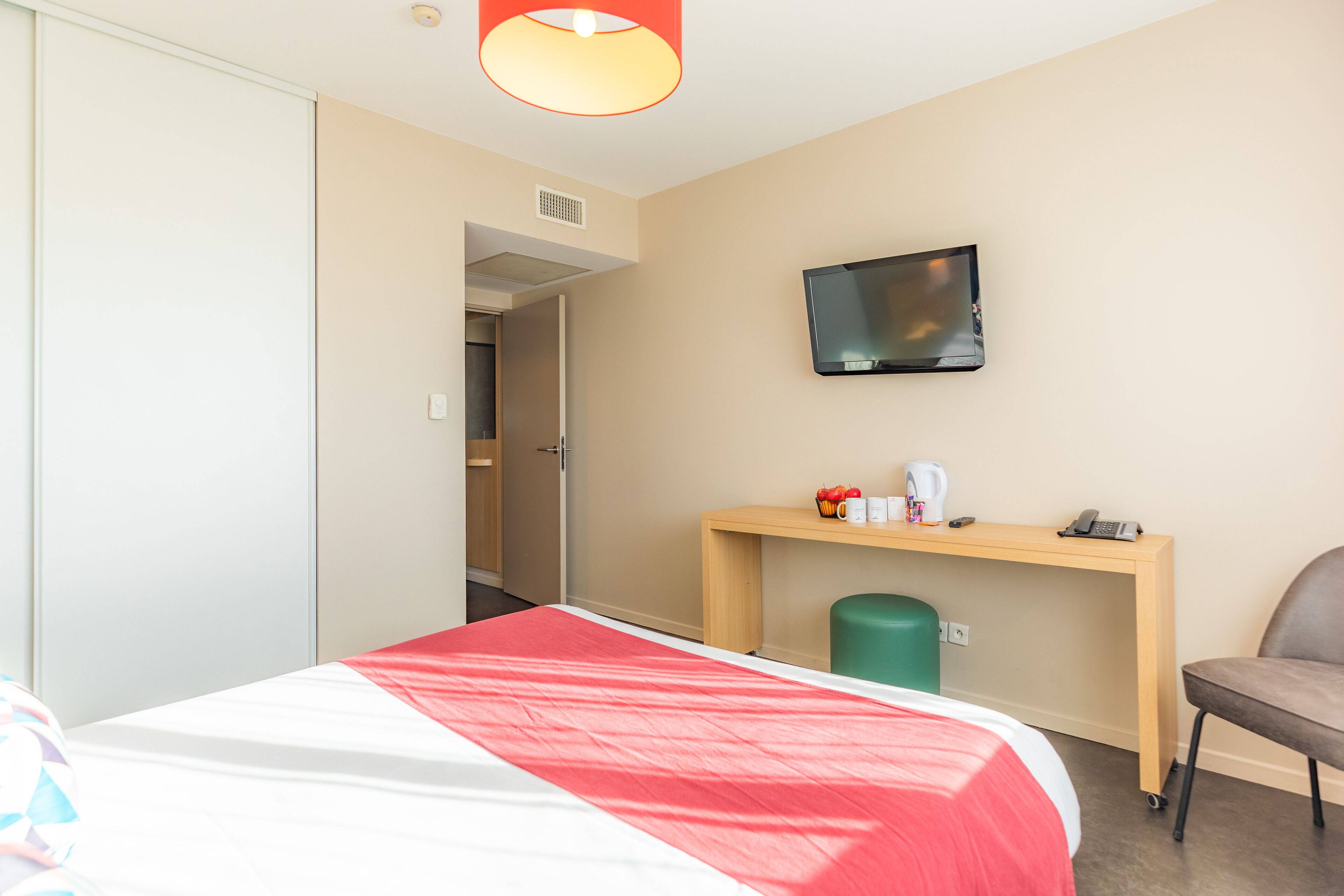 Ein Hotelzimmer mit einem Bett, einem Schreibtisch mit Fernseher, einem Sessel und einer grünen Mülltonne.
