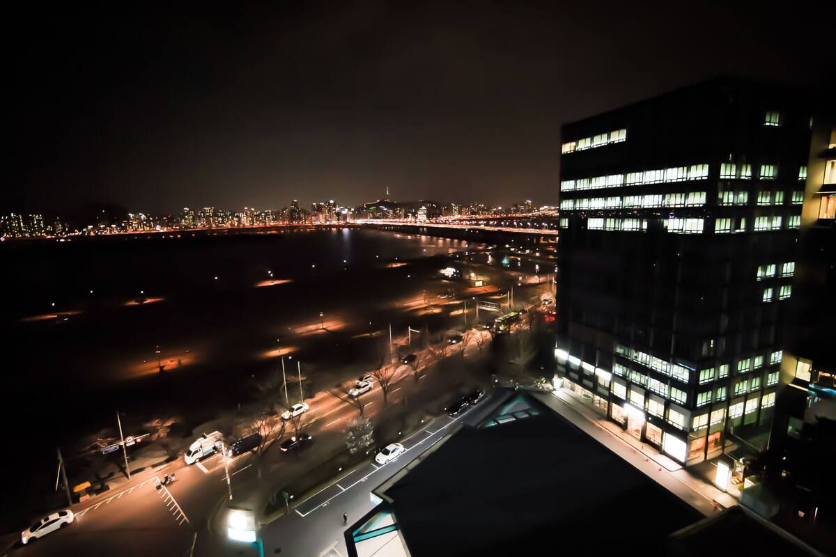 Un paisaje urbano nocturno con un puente, edificios iluminados y un estacionamiento.