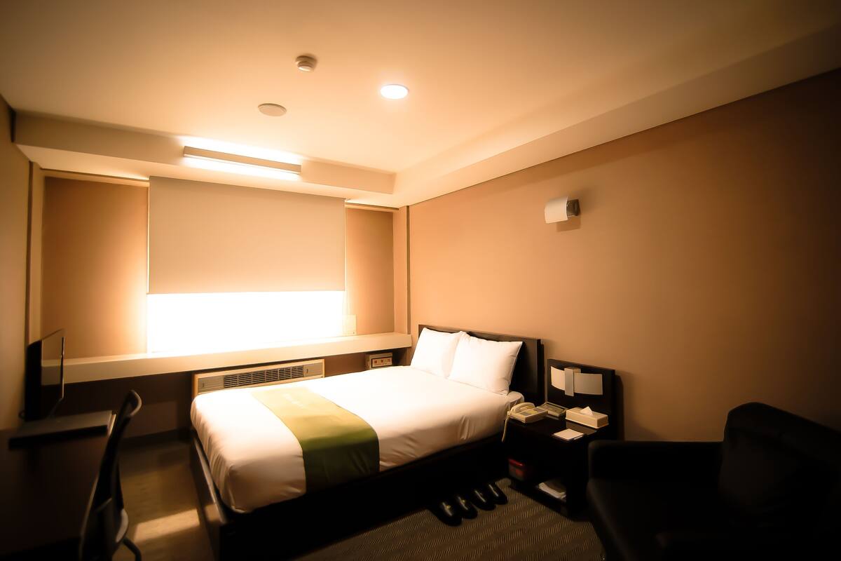 Una habitación de hotel con cama, escritorio, silla, mesita de noche y ventana.