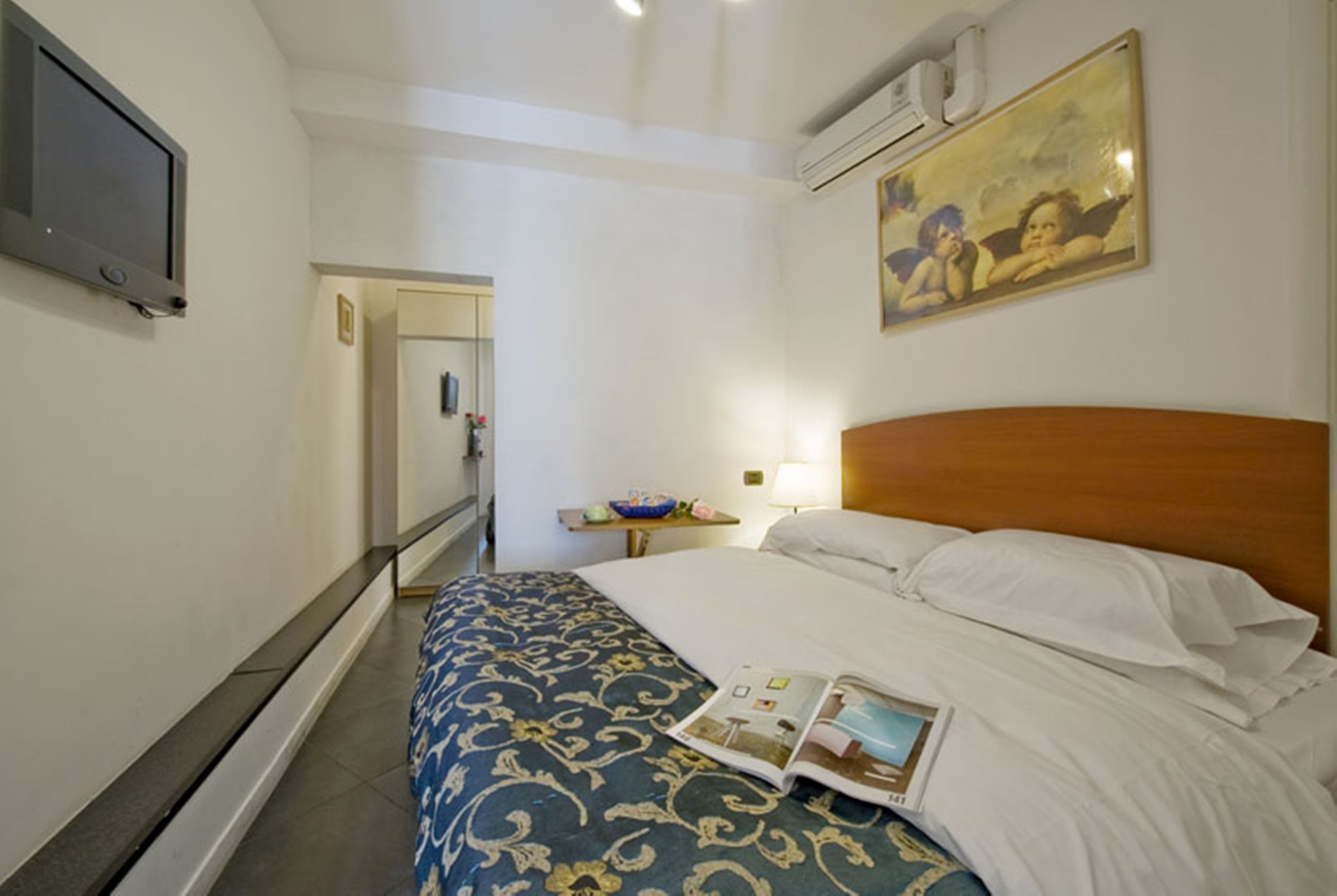 A hotel room with a bed, a bedside table with a lamp, a painting on the wall, and a television mounted on the wall.