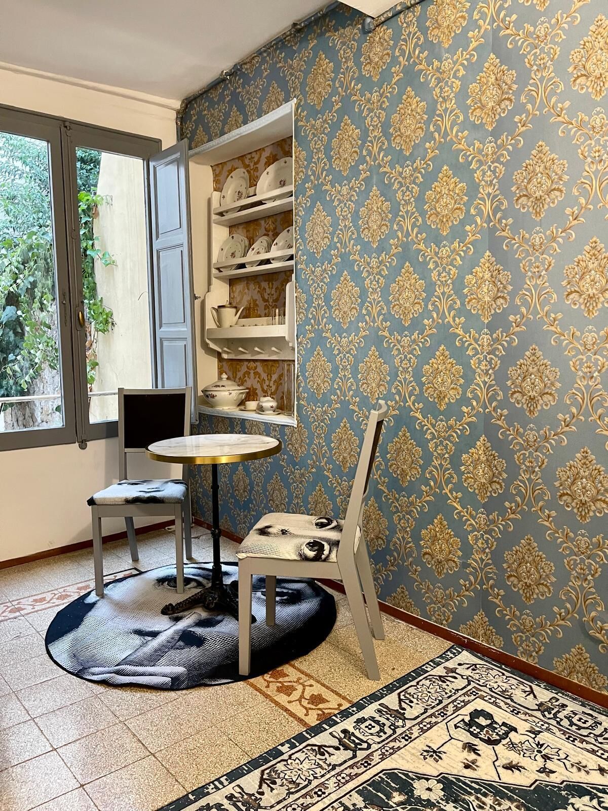 Une pièce avec un mur orné d’un motif bleu, une étagère blanche contenant de la vaisselle, une table ronde avec une chaise, et un tapis à motifs.