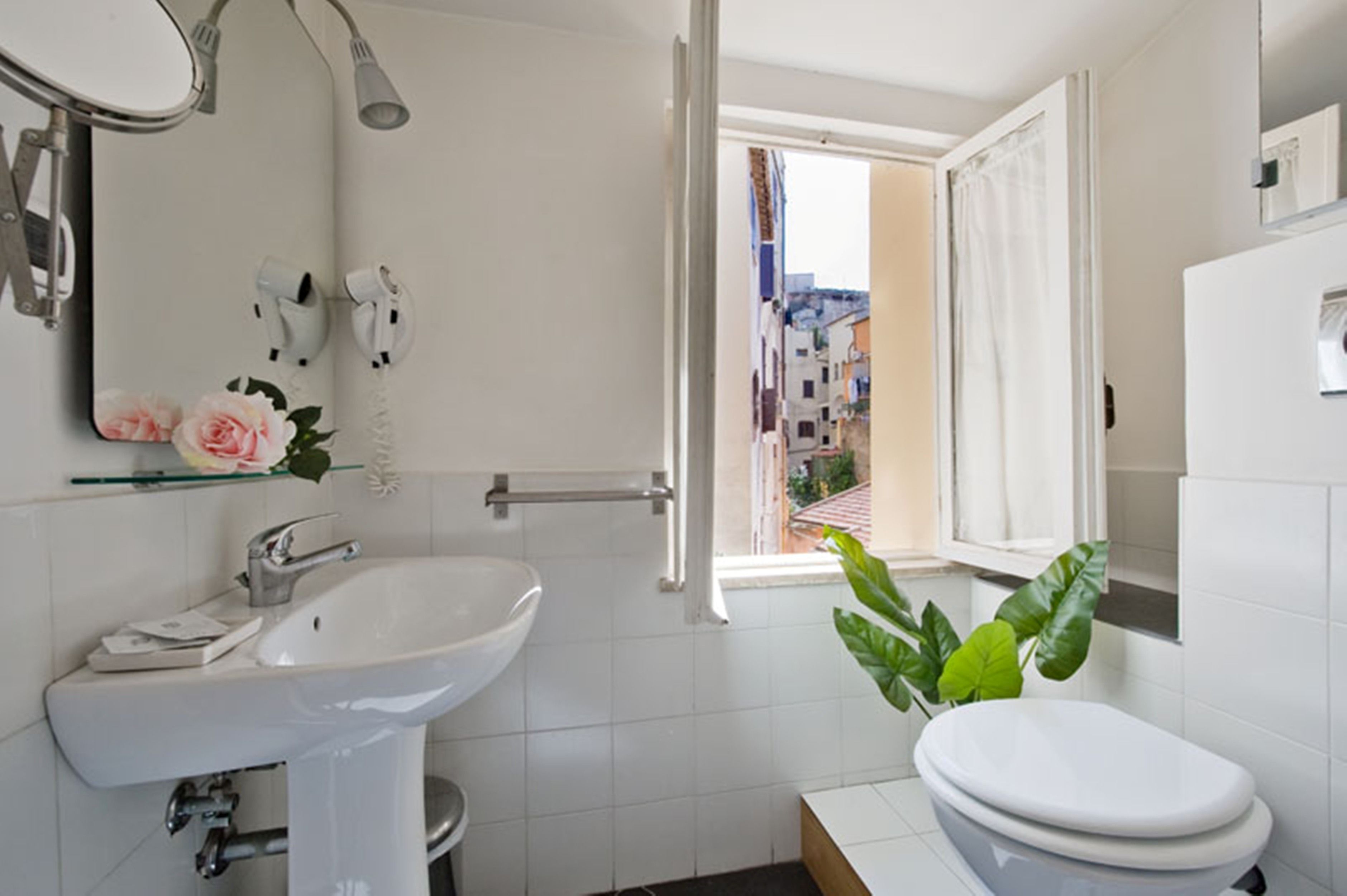 A modern bathroom with a white sink, toilet, and shower. A window provides a view of a cityscape.