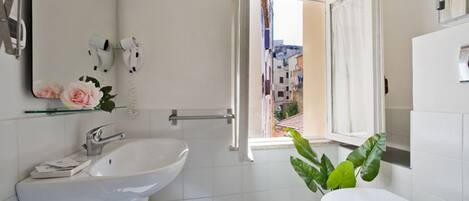 Un baño moderno con lavamanos, inodoro y ducha blancos. Una ventana ofrece vistas a un paisaje urbano.