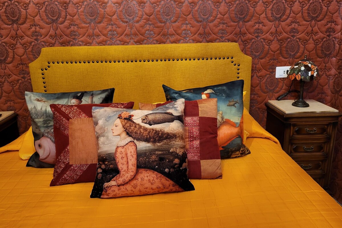 Un lit avec une tête de lit jaune, plusieurs coussins décoratifs et une table de chevet avec une lampe.