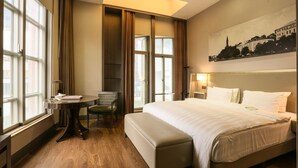 A hotel room with a large bed, a desk with a chair, a window with curtains, and a painting on the wall.