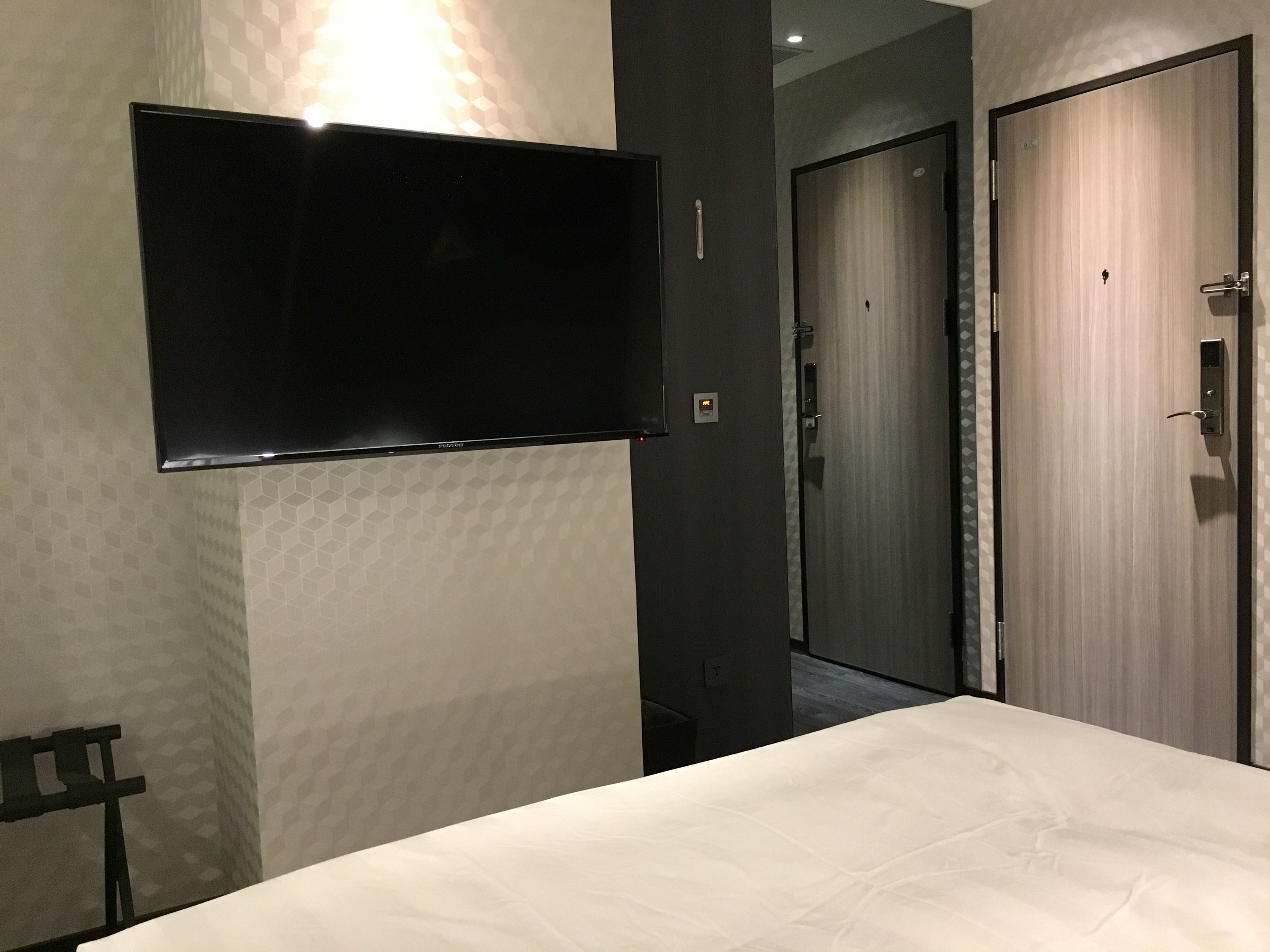 A hotel room with a bed, a flat-screen TV mounted on the wall, a door, and a wall with a geometric pattern.