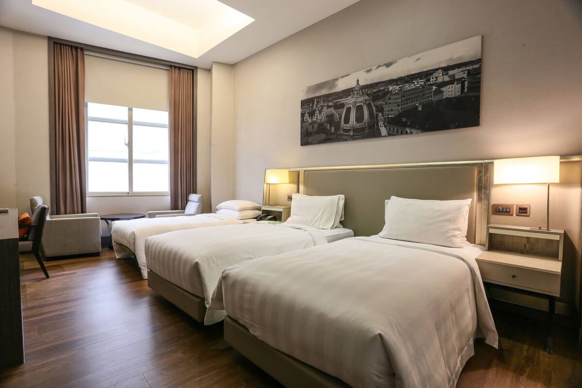Ein Hotelzimmer mit zwei Betten, einem Schreibtisch, einem Stuhl, einem Fenster mit Vorhängen und einem Wandbild.