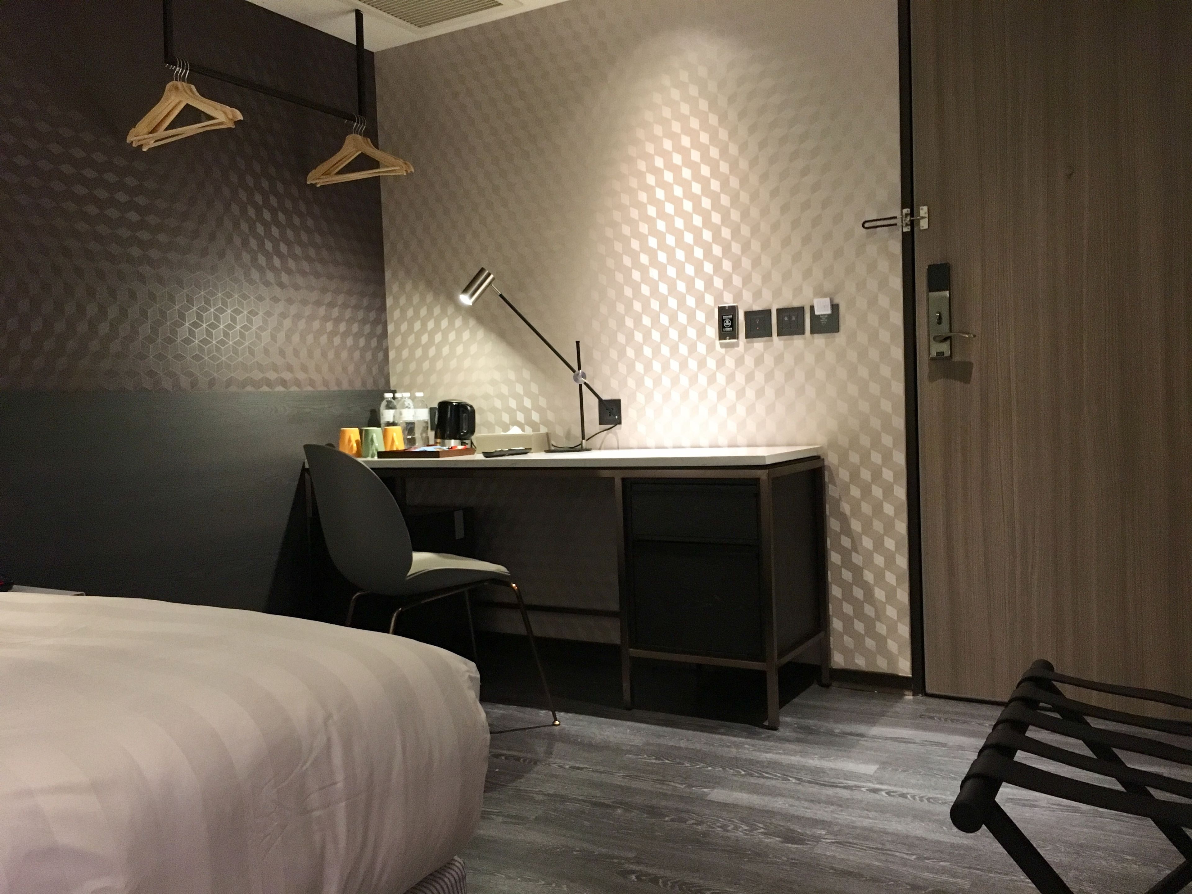 Una habitación de hotel moderna con cama, escritorio y silla. Hay perchas de madera en la pared, una lámpara de escritorio y una cafetera sobre el escritorio.