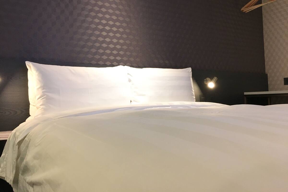 Ein ordentlich bezogenes Bett mit weißen Leinen in einem Zimmer mit gemustertem Wandpapier und einer Pendelleuchte.