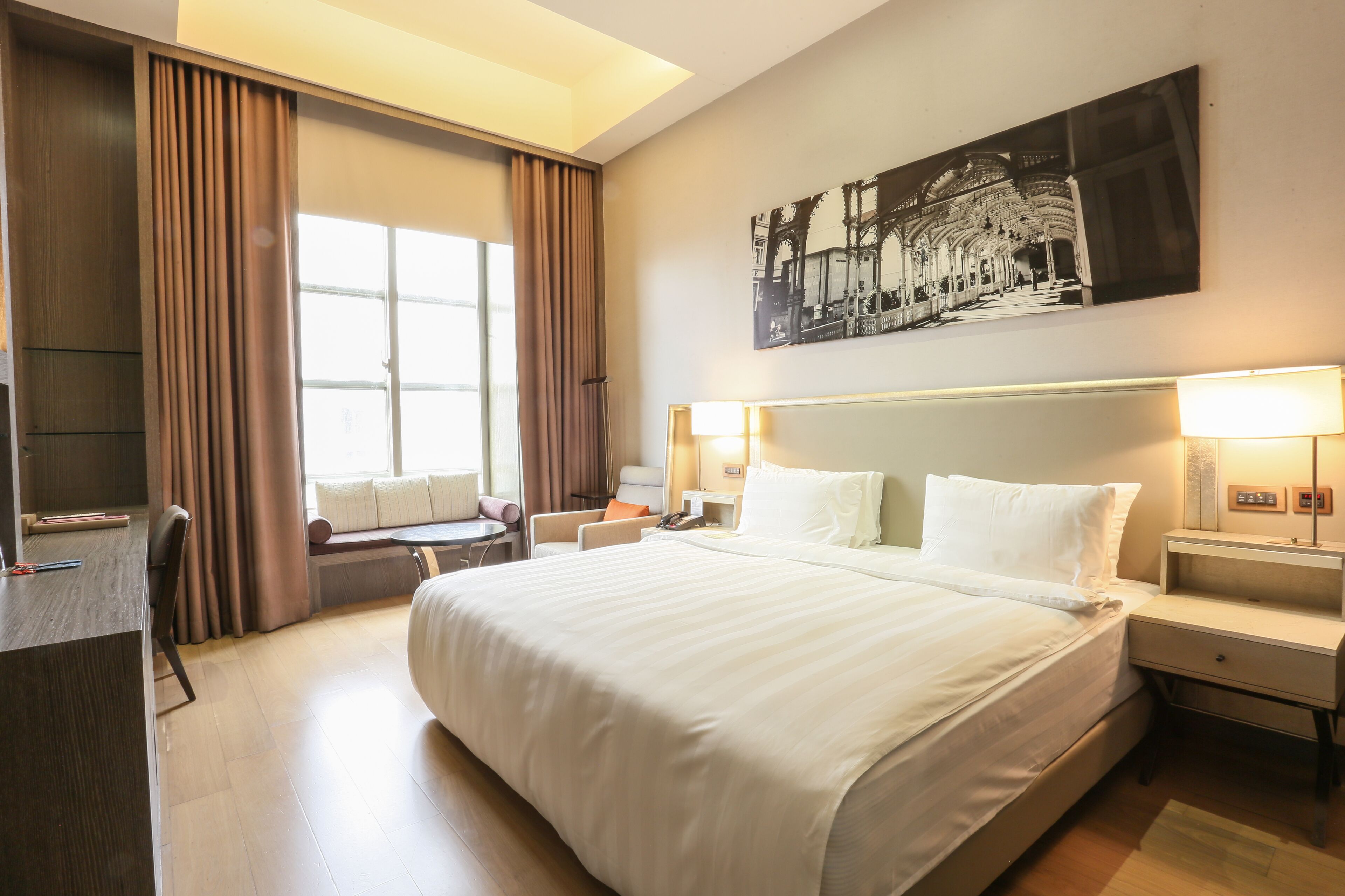A hotel room with a large bed, a desk, a chair, a window with curtains, and a wall-mounted artwork.