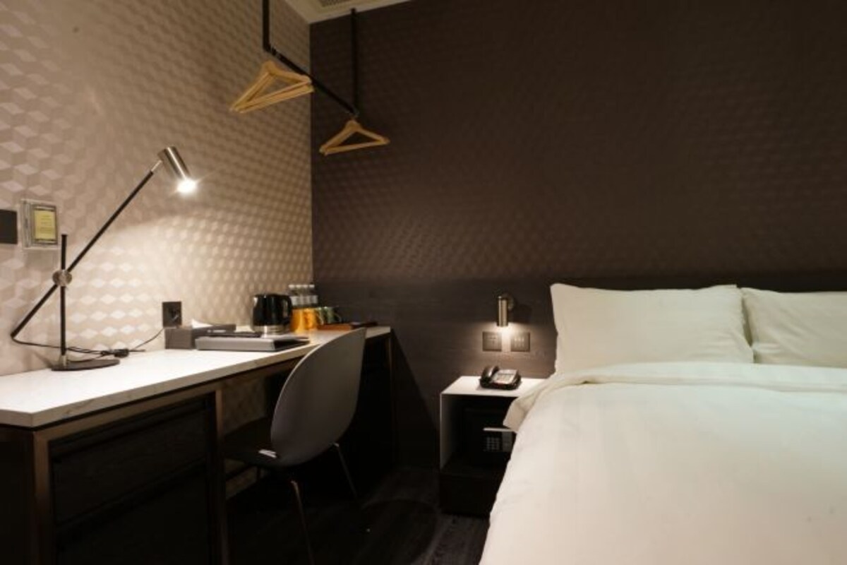 Een hotelkamer met een bureau, stoel, bed, nachtkastje, lamp en hanglamp.