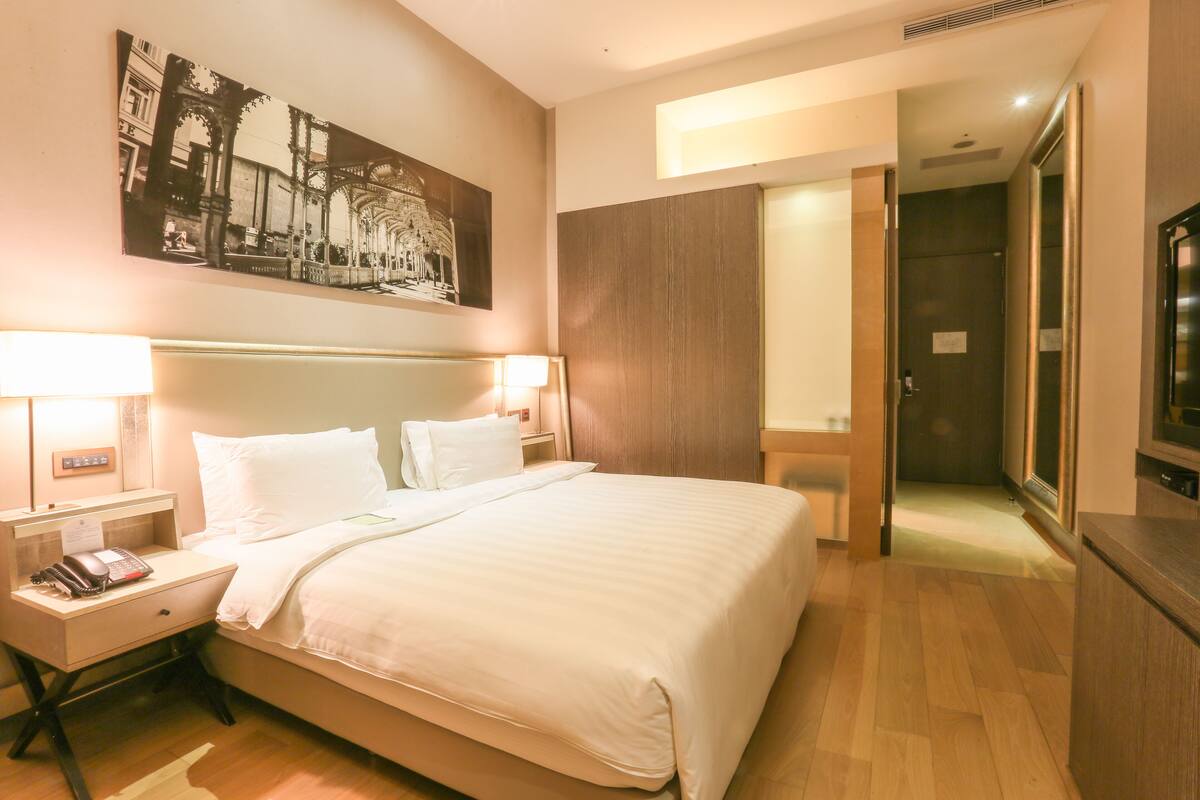 Ein Hotelzimmer mit einem großen Bett, Nachttischlampen, einem Telefon, einem Gemälde an der Wand und einem Fernseher auf einer Holzkommode.