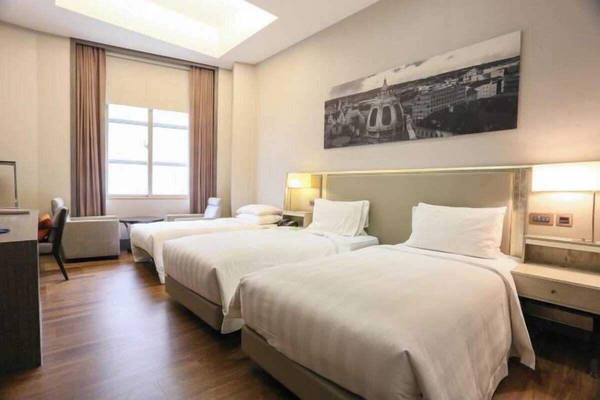 Ein Hotelzimmer mit zwei Betten, einem Schreibtisch, einem Stuhl und einem großen Fenster mit Vorhängen.