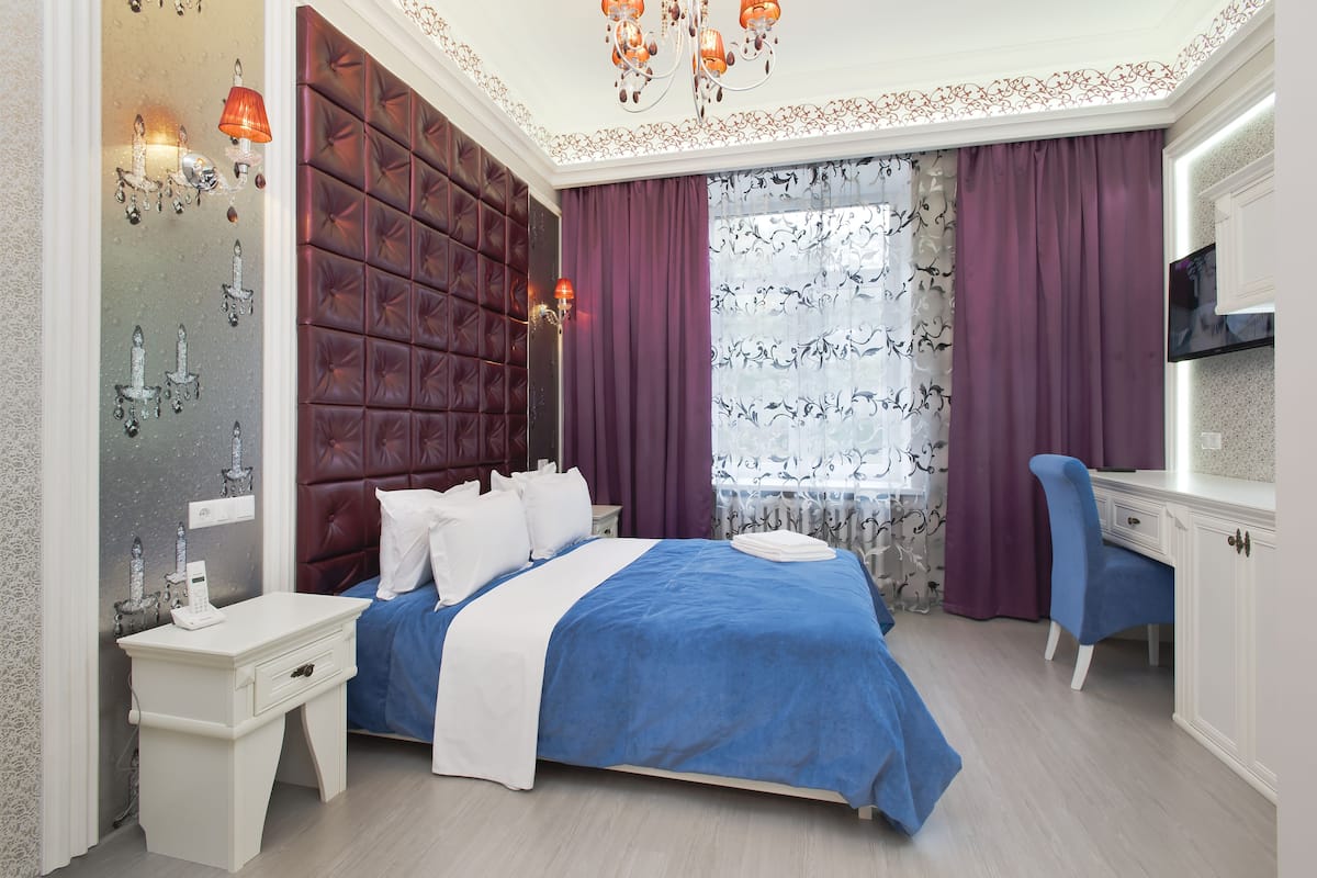 A bedroom with a tufted headboard, a bed with blue and white bedding, a bedside table with a lamp, a desk with a chair, and a TV on the wall.