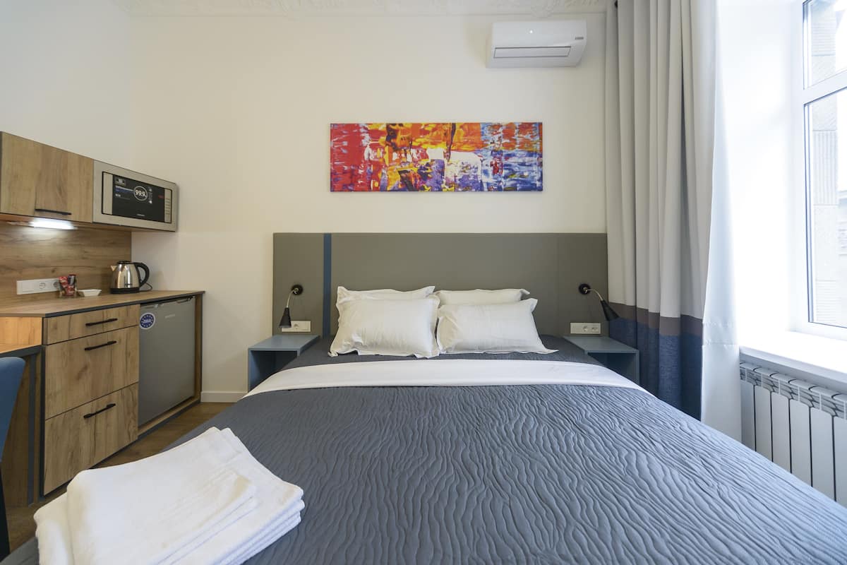 A hotel room with a bed, a desk with a kettle, a microwave, and a colorful abstract painting on the wall.