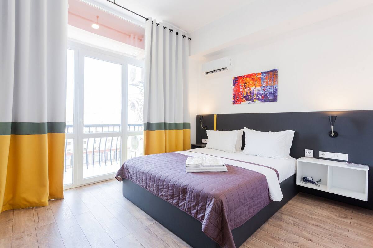 A hotel room with a large bed, a bedside table, a wall-mounted air conditioner, and a window with curtains.