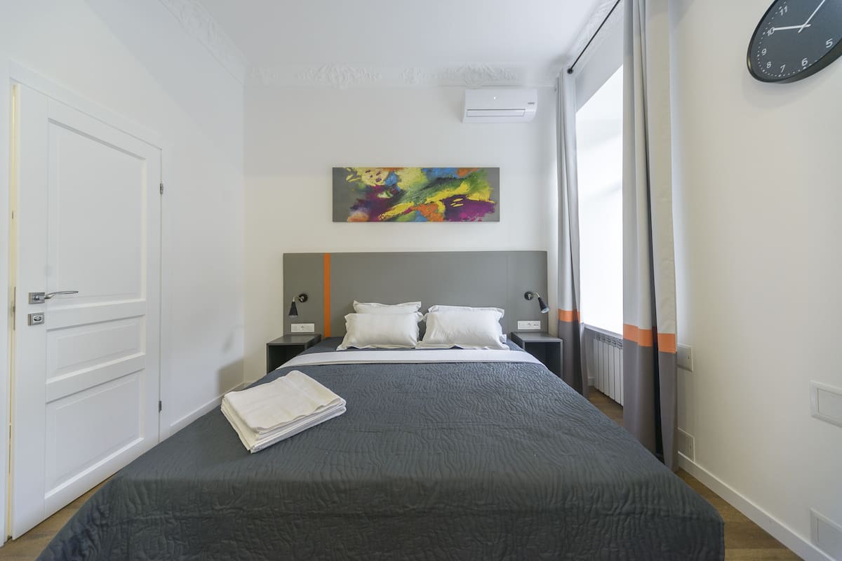 A hotel room with a bed, a colorful abstract painting, a clock, and a window with curtains.