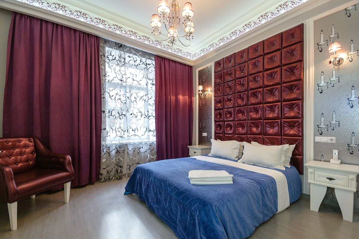 A bedroom with a large bed, a red upholstered headboard, a chandelier, and a red leather armchair.