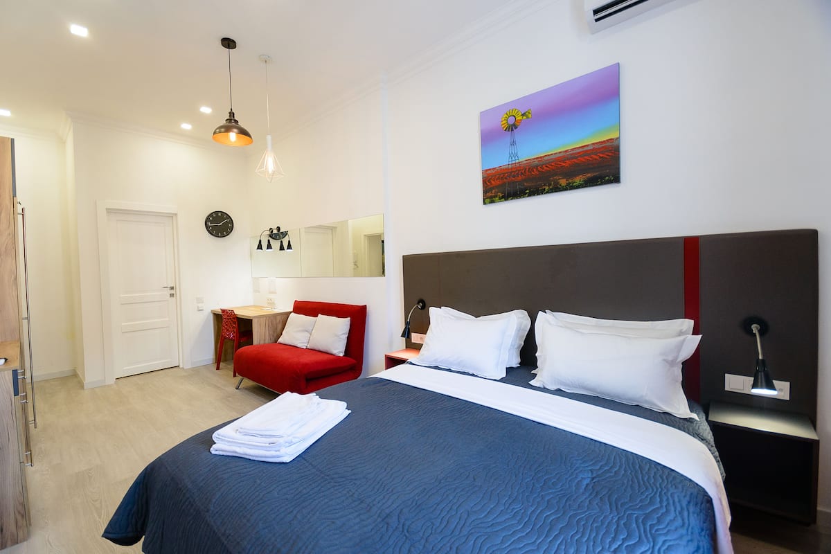 A hotel room with a large bed, a red chair, a desk, a mirror, and a painting on the wall.