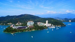 An aerial view of a coastal city with a prominent building complex, a small island with boats, and lush green hills in the background. - Tanxiang Resort Hotel Sun Moon Lake (Yuchi)