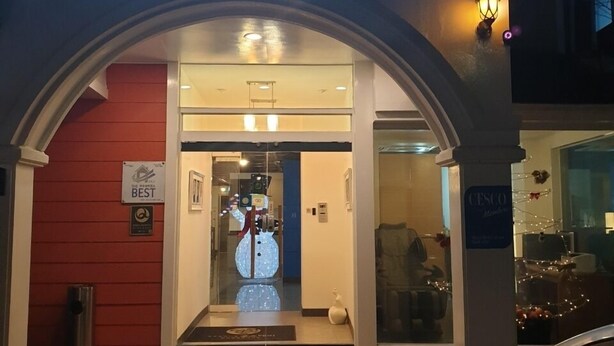 The entrance of a hotel with a neon sign above the door, a red wall on the left, and a car parked outside.