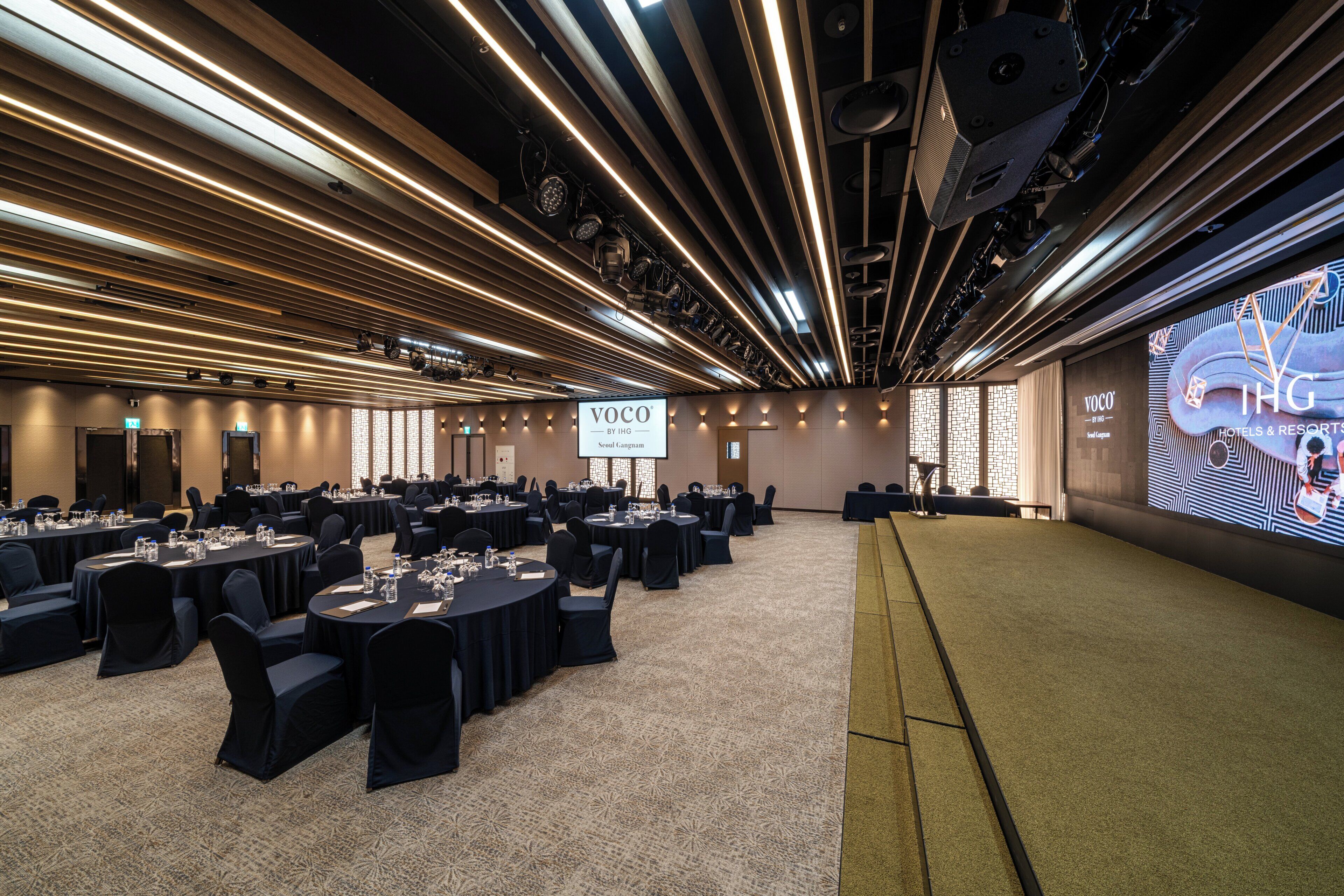 A conference room with multiple tables, chairs, and a large screen displaying 'Voco by IHG Seoul Gangnam'.