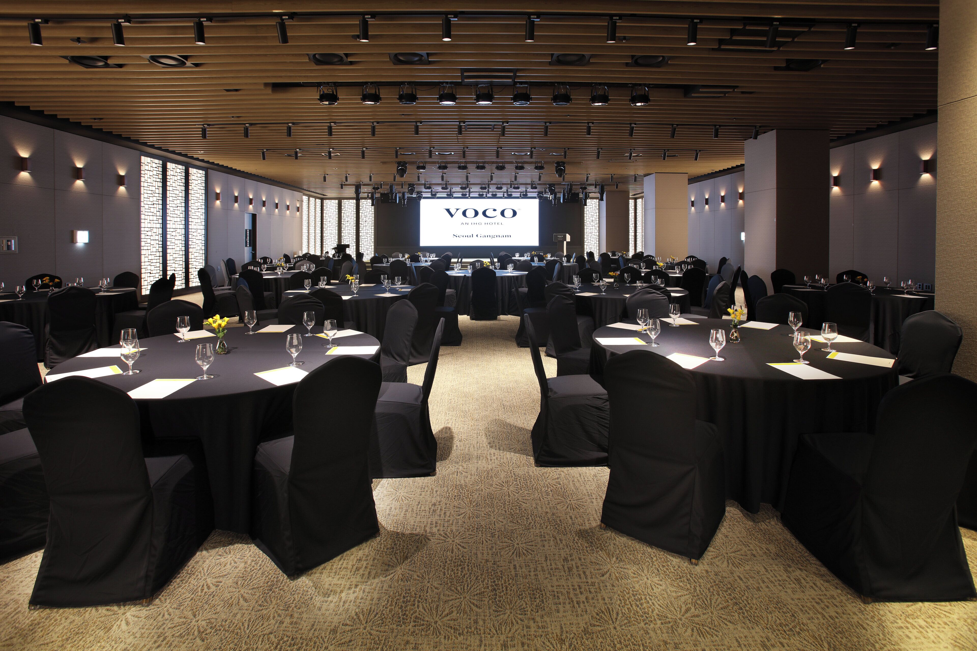 Un salón de conferencias con varias mesas redondas, sillas negras y una gran pantalla mostrando 'VOCO AN THE HOTEL Seoul Gangnam'.