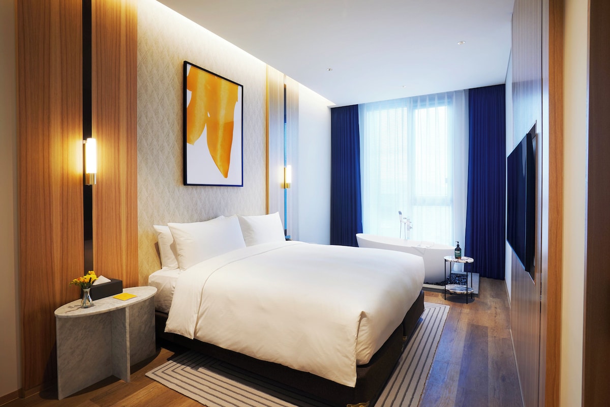 A modern hotel room with a large bed, a bedside table with a vase of flowers, a large window with curtains, and a wall-mounted artwork.