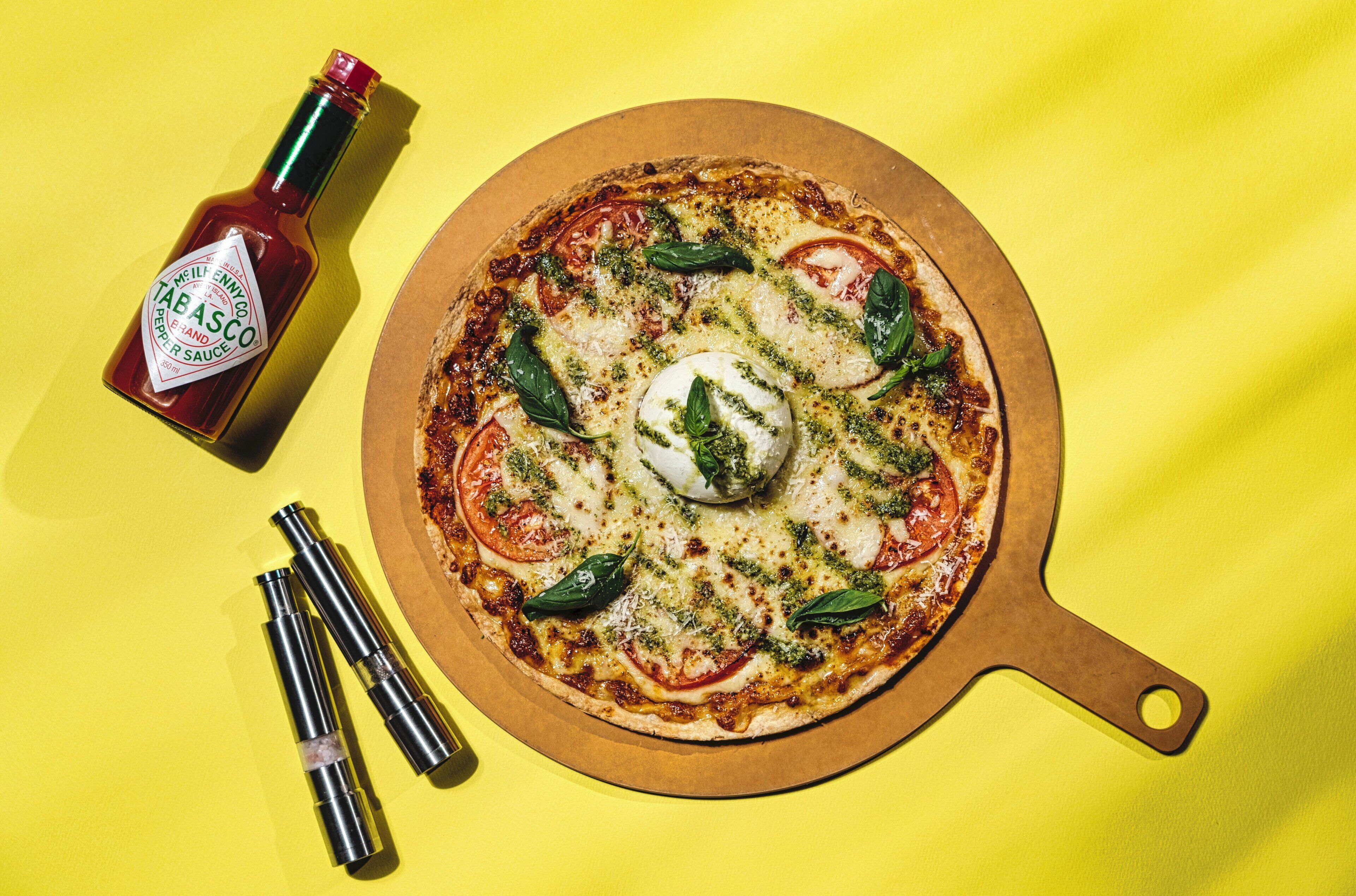 Pizza con tomate, albahaca y mozzarella sobre una tabla de madera, una botella de Tabasco y dos saleros sobre un fondo amarillo.