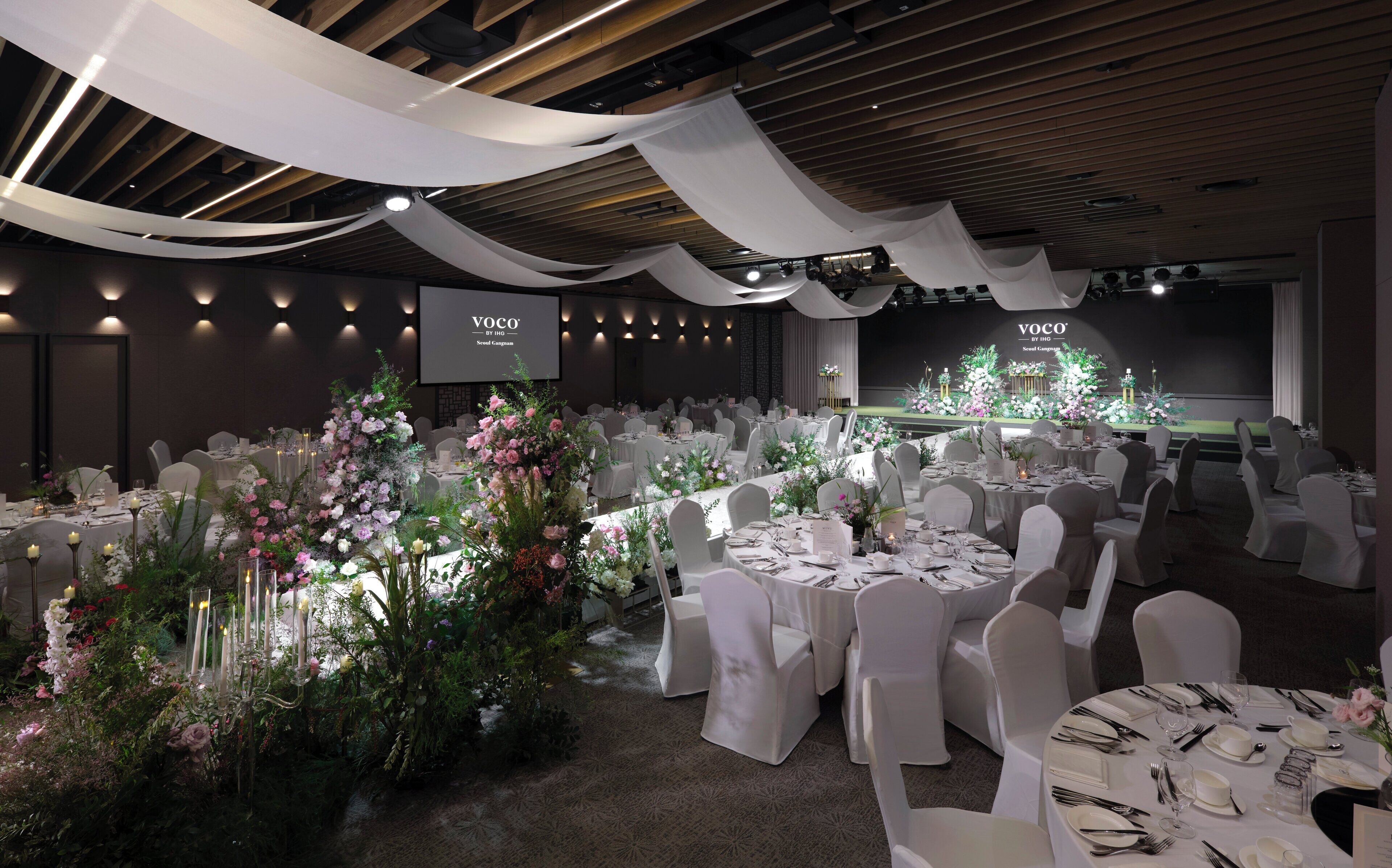 Un salón de banquetes con mesas redondas, sillas blancas y centros de mesa con flores.