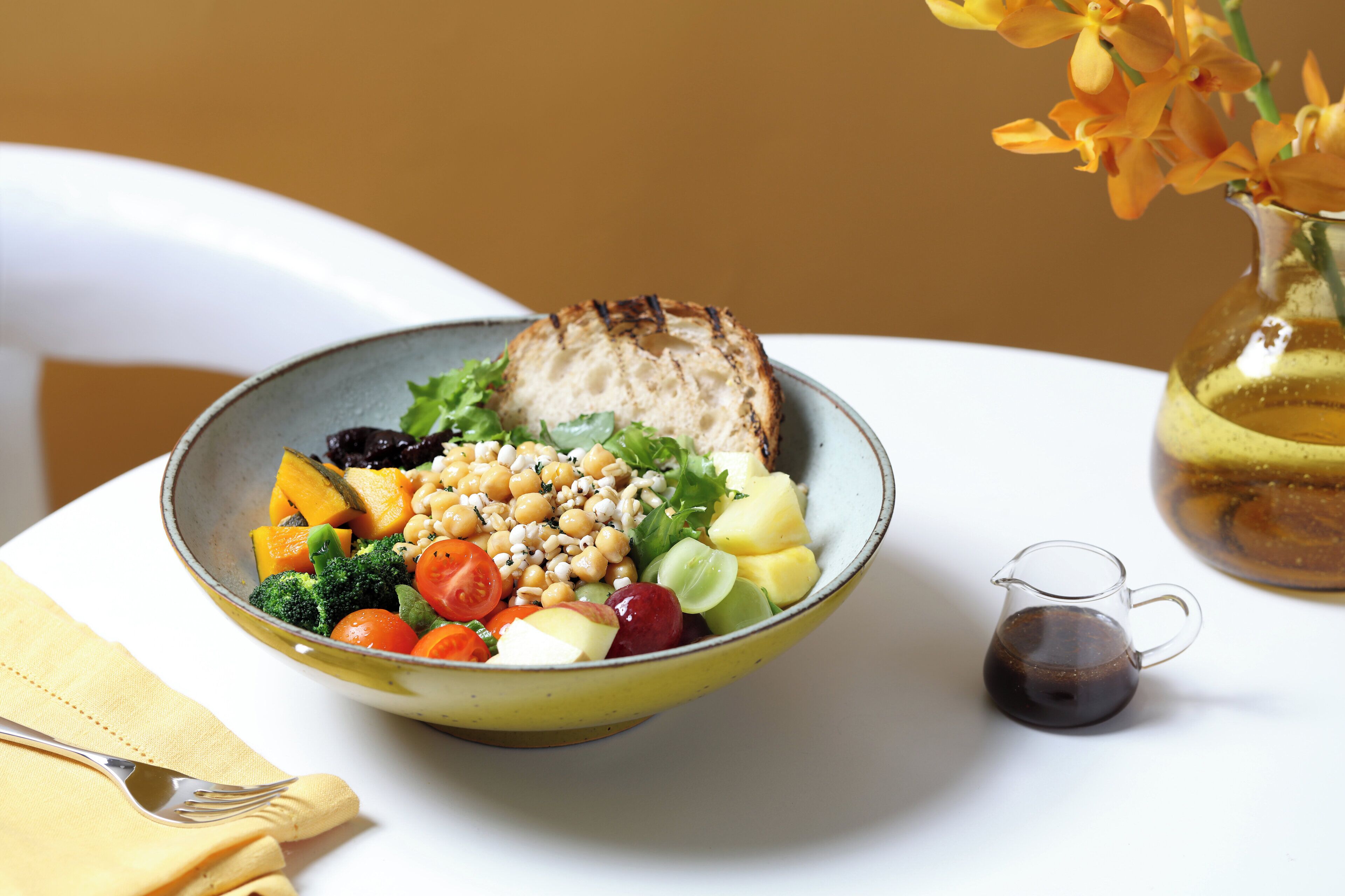 A bowl of salad with various vegetables and chickpeas, a small pitcher of sauce, and a vase with flowers on a table.