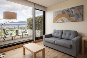 A modern living room with a grey sofa, a wooden coffee table, and a large window offering a city view.
