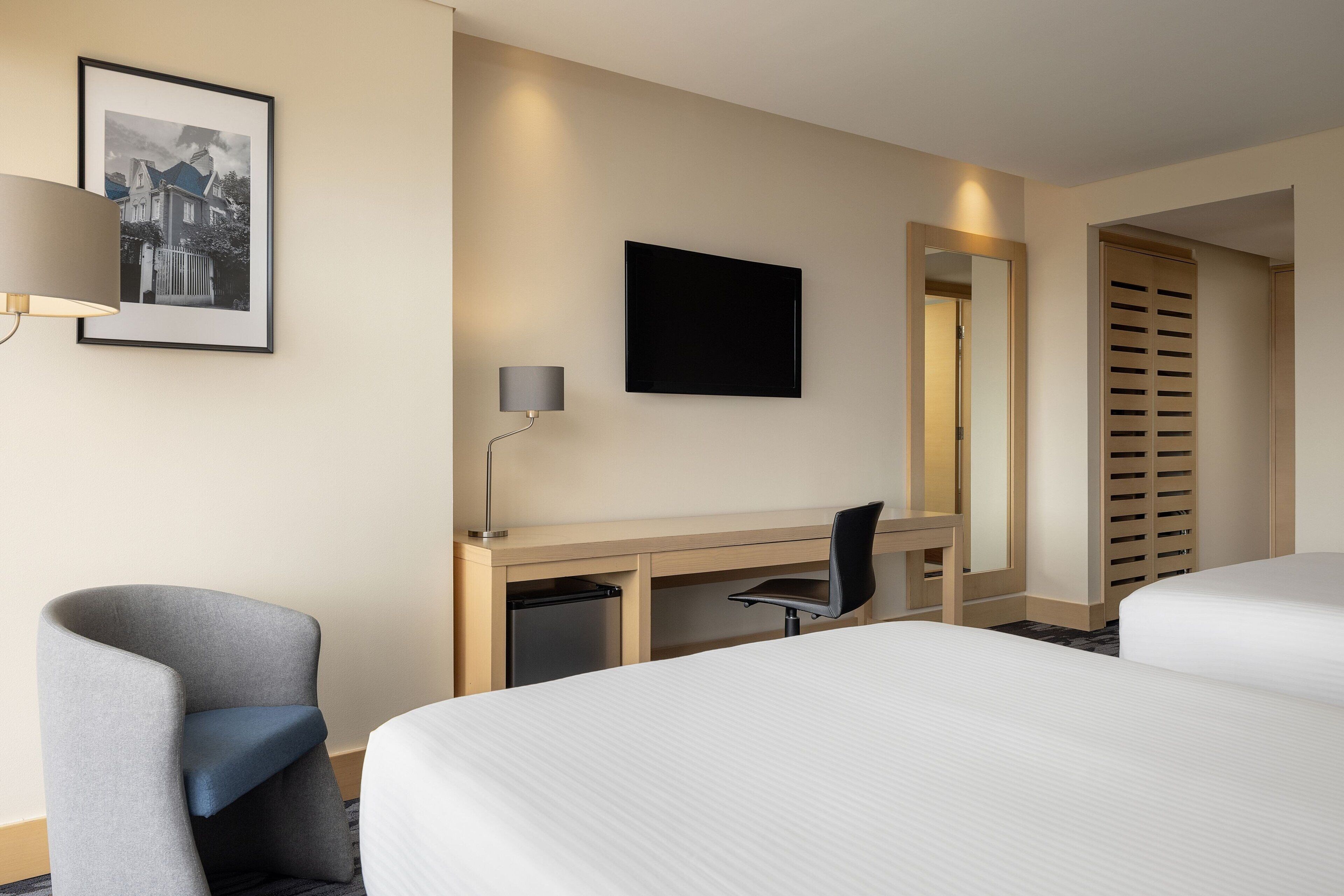 Une chambre d’hôtel avec deux lits, un bureau, une chaise, une télévision, une lampe et un tableau encadré au mur.